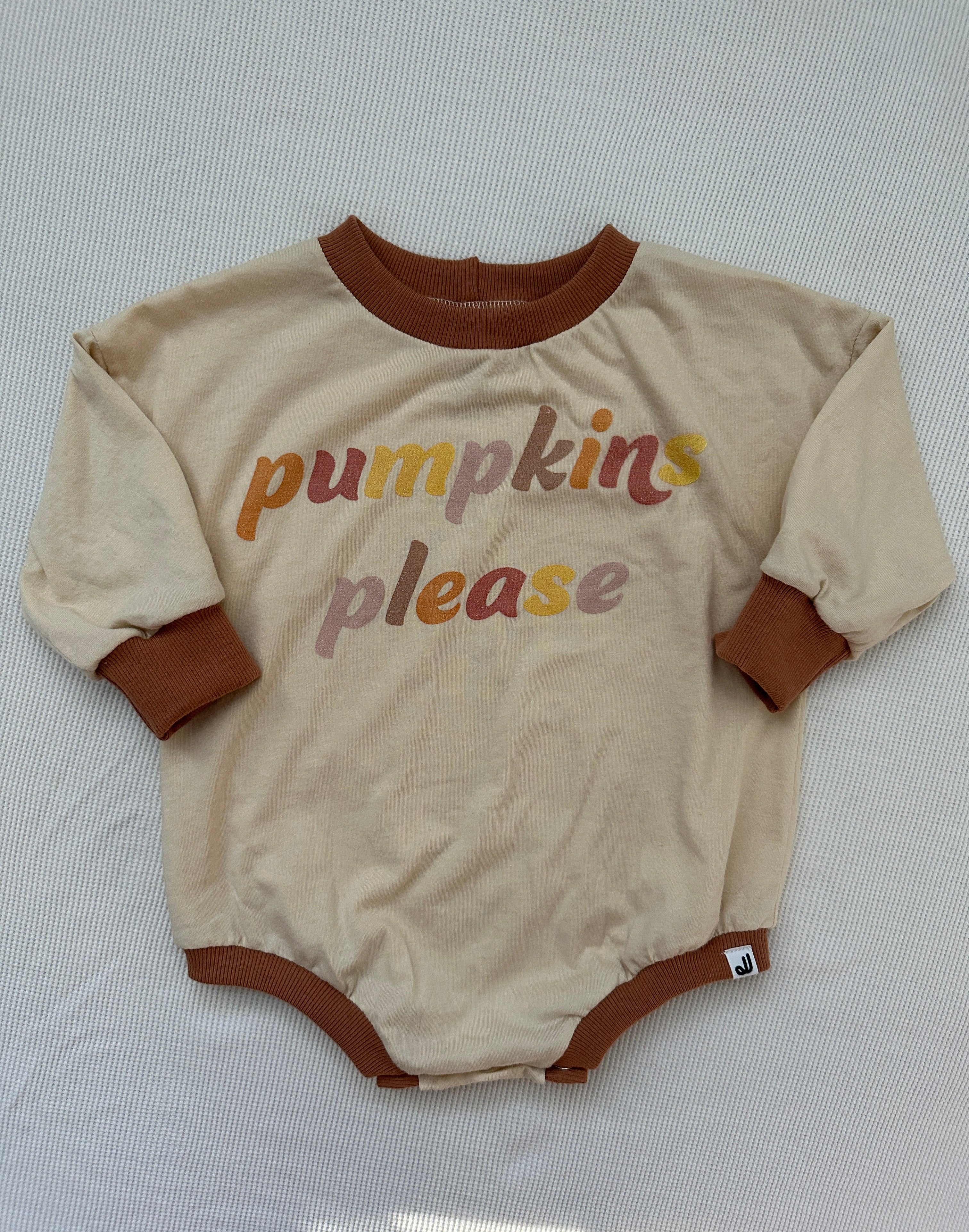A long sleeve romper with 'PUMPKINS PLEASE' printed in orange and brown colors, laid flat on a white surface.